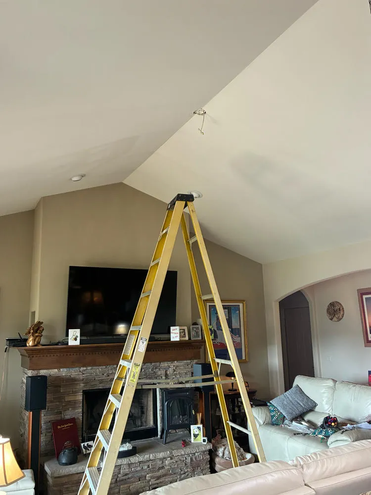 A tall yellow A frame ladder standing in a living room, positioned under exposed ceiling wires for light installation